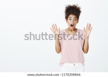 Portrait of surprised and amazed beautiful female with combed curly hair, raising spread palms and gasping, dropping jaw from excitement, seeing something unexpected, standing against gray background