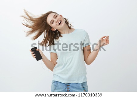 Music is great emotion booster. Portrait of charming joyful and emotive woman jumping happily waving hair and smiling from joy listening music in earphones holding smartphone posing against grey wall