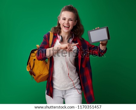 smiling young student woman in a red shirt with backpack and headphones pointing at tablet PC blank screen on green background