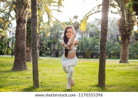 Young athletic woman in white sports outfit performing Yoga Eagle pose outdoor, in park meadow.