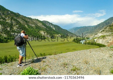 A man photographer is setting up a camera standing on a tripod on top of a rock in the mountains of the Altai makes a photo of a turquoise river of a reel and mountains with clear blue sky and clouds