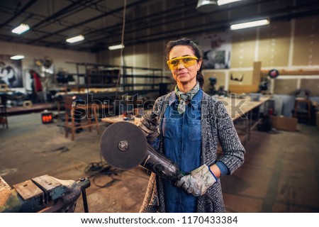 Picture of professional middle age female carpenter standing in her workshop and holding electric drill. Looking at camera and smiling.