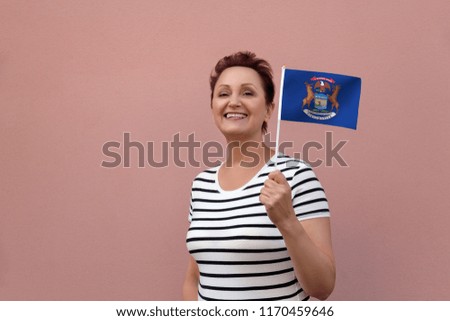 Michigan flag. Woman holding Michigan state flag. Nice portrait of middle aged lady 40 50 years old with a state flag over pink wall on the street outdoors.