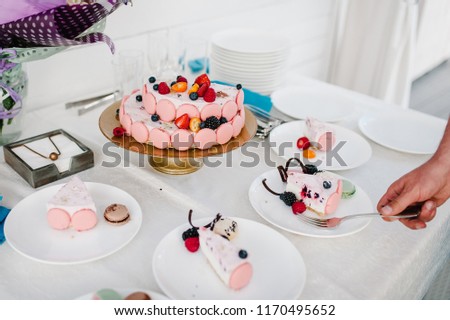 A pieces of Birthday pink cake decorated with fresh berries: strawberries, raspberries, currants, blackberries, blueberries and macarons in plate on table for holiday. Man's hands take fork.