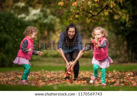 Mother and identical twins having fun under tree with autumn leaves in the park, blond cute curly girls, happy family, beautiful girls in pink jackets, young family, healthy lifestyle, colorful season