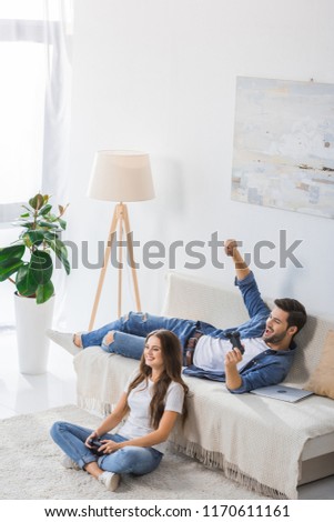 high angle view of happy young man with joystick gesturing by hand and celebrating victory while his girlfriend sitting on floor at home