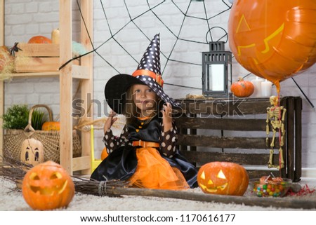 Happy Halloween. A little beautiful girl in a witch costume celebrates a home in an interior with pumpkins