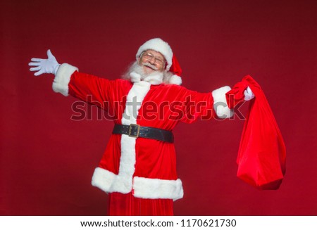 Christmas. Kind smiling Santa Claus spread his hands to the sides. In one hand he is holding a red bag with gifts. Isolated on red background.