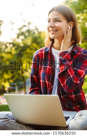 Picture of young amazing lady student sitting in the park using laptop computer listening music with earphones.
