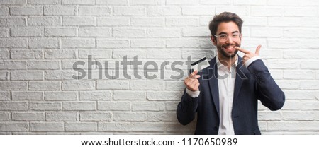 Young business man wearing a suit against a white bricks wall smiles, pointing mouth, concept of perfect teeth, white teeth, has a cheerful and jovial attitude