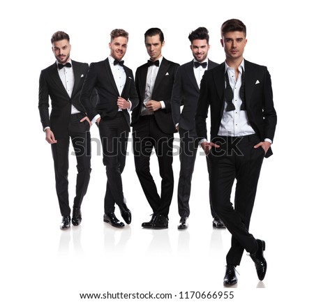 five groomsmen in black tuxedoes with leader with undone collar standing in front cross-legged, on white background, full body picture