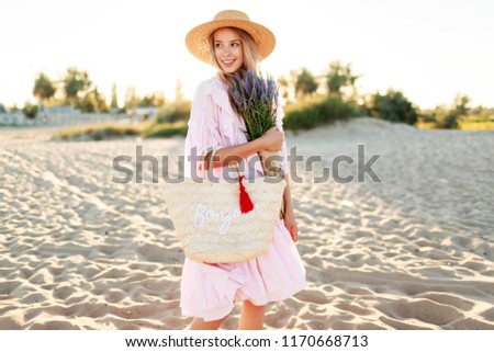 Elegant white woman  with flowers in hands walking on sunny beach . Warm sunset colors. Straw hat.  
Pink dress. Holidays and vacation mood. 