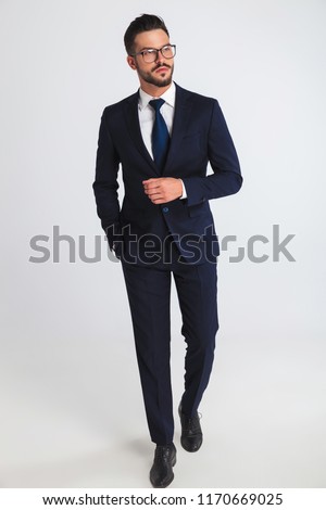 relaxed businessman wearing glasses and navy suit walking and looking to side on light grey background, full body picture