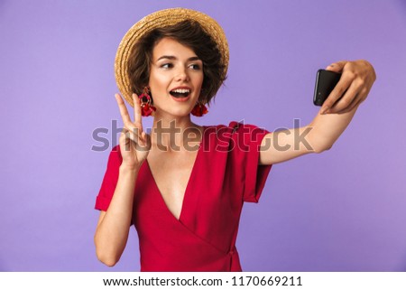 Happy elegant woman in dress and straw hat making selfie on smartphone while showing peace gesture over violet background