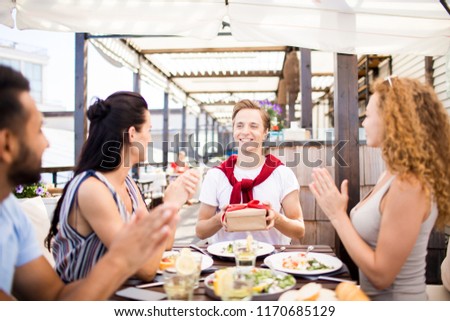 Portrait of group of friends celebrating birthday in outdoor cafe, focus on happy man holding gift box with red ribbon, copy space