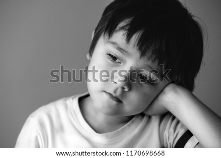 Black and white photo of Closeup beautiful innocent eyes of little boy laying down and looking out with thinking of some thing,Lonely kid sitting alone putting head down on his hand with bored face,