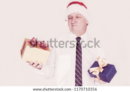 Pensive grimacing mature businessman choosing gift and looking with doubt at gift box. Puzzled man in Santa hat holding Christmas presents. Gift store concept
