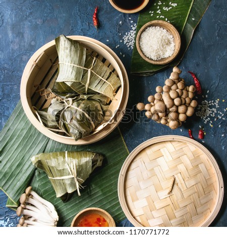 Asian rice piramidal steamed dumplings from rice tapioca flour with meat filling in banana leaves served in bamboo steamer. Ingredients, sauces above. Blue texture background. Top view. Square image