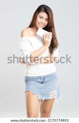 Studio shot portrait of young beautiful Asian girl standing and hold smartphone in hand with smiling face on light grey background