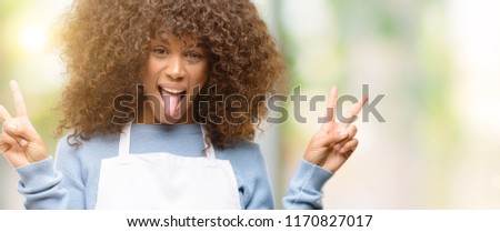 African american shop owner woman wearing an apron looking at camera showing tongue and making victory sign with fingers