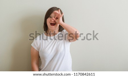 Down syndrome woman standing over wall doing ok gesture shocked with surprised face, eye looking through fingers. Unbelieving expression.