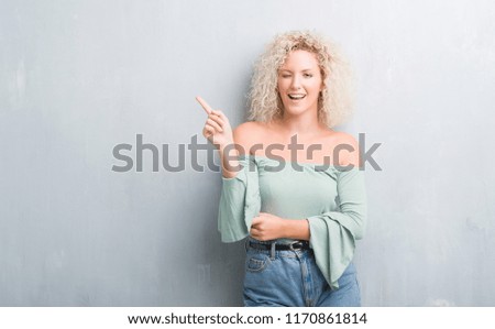 Young blonde woman with curly hair over grunge grey background with a big smile on face, pointing with hand and finger to the side looking at the camera.