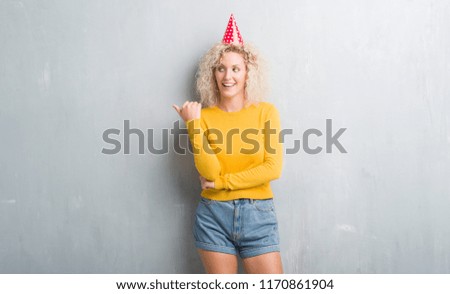 Young blonde woman over grunge grey wall wearing birthday hat smiling with happy face looking and pointing to the side with thumb up.
