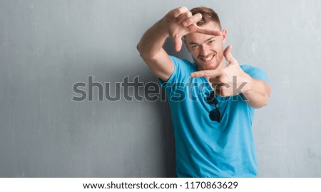 Young redhead man over grey grunge wall wearing casual outfit smiling making frame with hands and fingers with happy face. Creativity and photography concept.