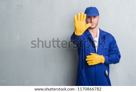 Young caucasian man over grey grunge wall wearing mechanic uniform with open hand doing stop sign with serious and confident expression, defense gesture