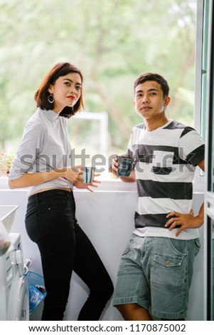 A portrait of a young Malay Asian couple enjoying a hot drink and talking to one another fondly. They are sharing a tender moment together during the day by the window balcony. 