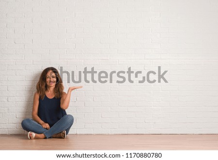 Middle age hispanic woman sitting on the floor over white brick wall smiling cheerful presenting and pointing with palm of hand looking at the camera.