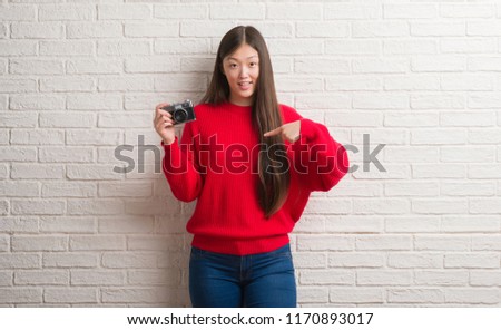 Young Chinese woman over brick wall holding vintage camera with surprise face pointing finger to himself