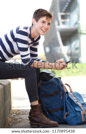 Side portrait of happy young woman sitting outdoors with mobile phone and bag