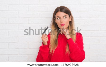 Young adult woman over white brick wall holding credit card serious face thinking about question, very confused idea