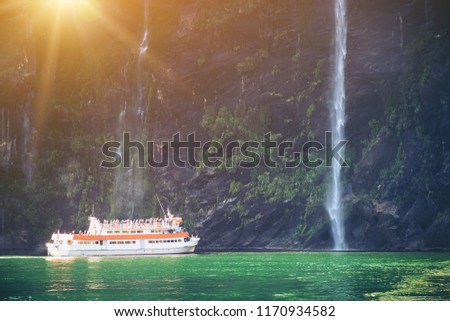 Sightseeing boat carrying tourist people approaches great waterfall in Milford Sound. Beautiful scenic cruise through Fiordland National Park in South Island of New Zealand.