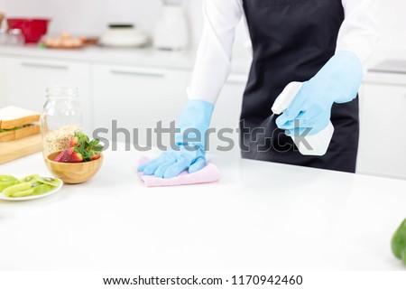 Attractive beautiful woman or housewife is wiping, cleaning and spaying antiseptic or disinfectant on table at kitchen. Cleaning can kill germ, bacteria. Charming beautiful girl wear gloves, apron. Royalty-Free Stock Photo #1170942460