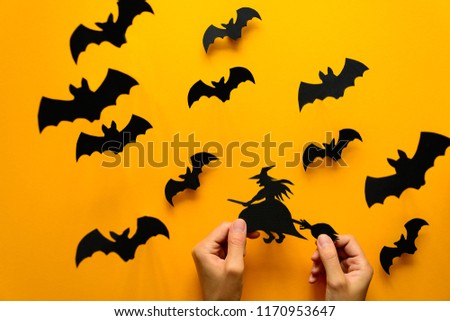 Woman's hand holding witch on broom and black paper bats flying on yellow background. Halloween concept. Paper cut style. Top view