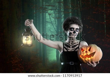 Witch with sugar skull makeup holding pumpkin at scary forest background. Copy space