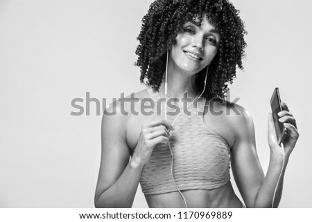 Black and white photo. Beautiful young and fit woman in headphones listening music from phone and smiling. Isolated on a yellow background in a pink sportswear