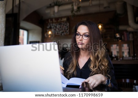 Woman in stylish glasses skilled copywriter working on web site via laptop computer while sitting in coffee shop.Female professional marketing writer preparing an article for publication using netbook