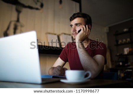 Handsome man freelance distance worker calling via mobile phone during job via laptop computer. Young male college student talking via cell telephone while sitting with netbook in modern coffee shop