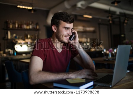 Smiling business man talking with client via mobile phone and reading information on laptop computer while sitting in coffee shop during work break. Happy hipster guy having smartphone conversation 