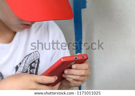 A teenager in a red baseball cap and white T-shirt is holding a red smartphone. Concept - game on a smartphone, virtual world, game addiction