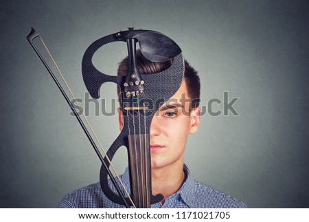 Young man in shirt covering face with violin in modern design looking at camera on gray background