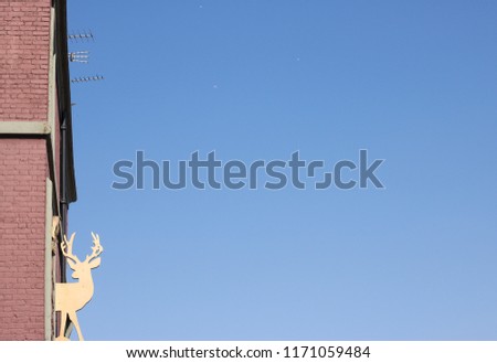 Image of a deer is attached to red brick wall of house. This decoration is high against the blue sky, flying airplanes, roof of city house and TV antennas. Concept: flight of imagination & fantasy.