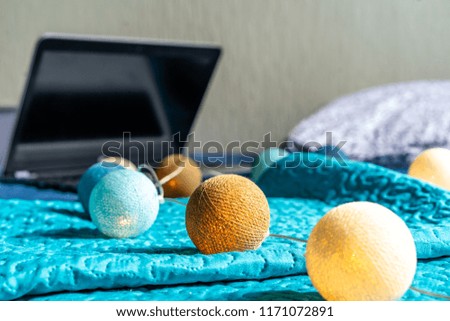 Grey Laptot Laying on Mattress with Turquoise Cover, Few Pillows and a White Background - with Christmas Decoration Lights on Bed
