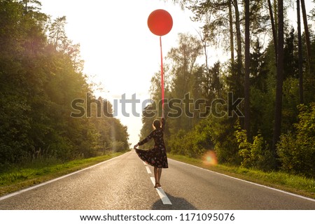 A young girl in a beautiful dress with flowers with a red balloon on a leash is walking along the road at sunset.
