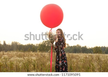 Young girl in a beautiful dress with flowers with a red balloon on a leash in the field at sunset.