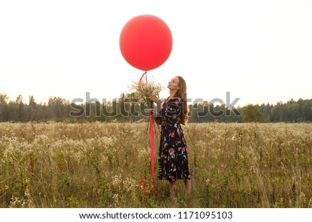 Young girl in a beautiful dress with flowers with a red balloon on a leash in the field at sunset.