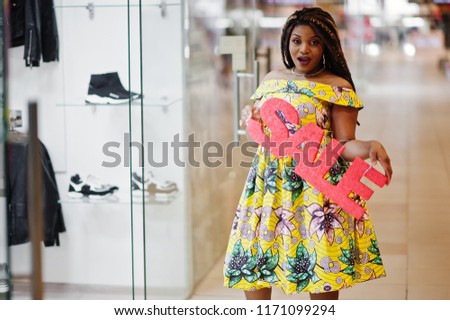 Cute small height african american girl with dreadlocks, wear at coloured yellow dress, holding sale sign letter on shopping center.